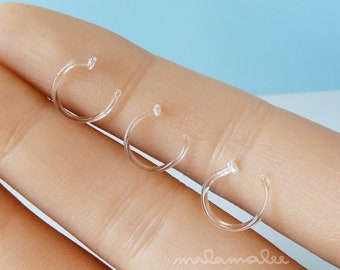 May include: Three clear, small, C-shaped nose rings are displayed. The rings are made of a transparent material and are designed to be nearly invisible when worn. The image is taken from a close-up perspective, highlighting the delicate design of the jewelry.