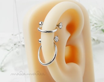 May include: A silver helix piercing with two clear gems and a silver hoop. The piercing is shown in a model's ear.