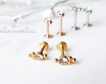 May include: Gold-toned lip piercing jewelry with a cluster of small beads and a clear gem. Several other pieces of jewelry are visible in the background, including rose gold and silver-toned options. The word "malamalee" is written in cursive.
