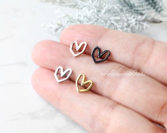 May include: Four small heart-shaped earrings in silver, gold, and black metal. The earrings are displayed on a hand.