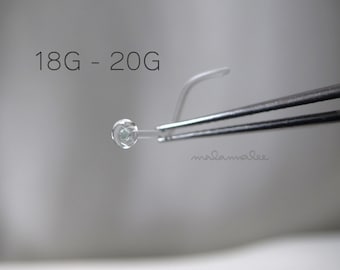 May include: A close-up of a clear glass bead with a small green center, held by a pair of tweezers. The text "18G - 20G" is visible above the bead.