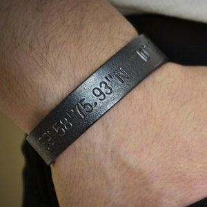 Puede incluir: Una pulsera de cuero negro con las coordenadas "43.68175.931'N" grabadas en ella.