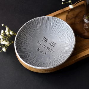 May include: A small, silver-colored dish with a textured surface and a wooden base. The dish is engraved with the date 06-19-2016 and the initials G & A. White flowers in a vase sit on a wooden tray.
