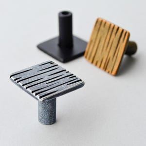 May include: A selection of cabinet knobs in different styles. One is black, square, and cylindrical. Another is silver with a textured, square face. The third knob has a square, wood-grain face. These knobs are suitable for adding a unique touch to furniture.