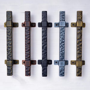 May include: Five different metal door pulls in various finishes. The pulls are all the same size and shape, with a hammered texture. The finishes include brass, bronze, black, silver, and antique gold.