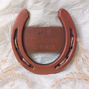 May include: A copper-colored horseshoe with a wooden plaque attached. The plaque has the text "IIII II CARL & GRACE 06-15-2016" engraved on it.