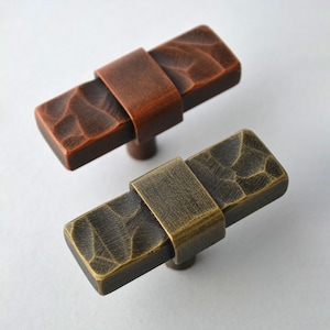 May include: Two rectangular cabinet pulls with textured surfaces. One is a reddish-brown color, and the other is a bronze-gold color. Each pull has a band around the center and a small, round base.