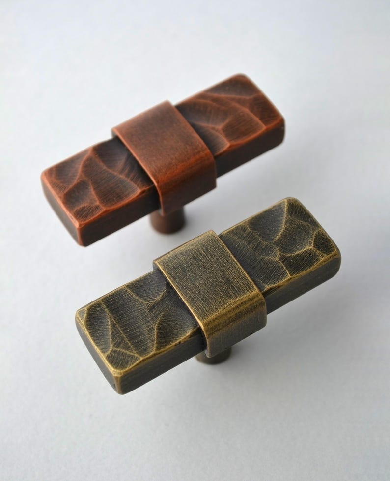 May include: Two rectangular cabinet pulls with textured surfaces. One is a reddish-brown color, and the other is a bronze-gold color. Each pull has a band around the center and a small, round base.