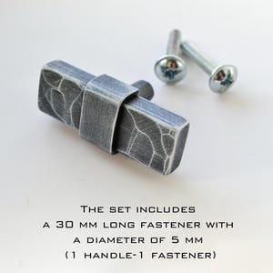 May include: A set of gray metal cabinet hardware. The set includes a 30 mm long fastener with a 5 mm diameter. The handle has a textured surface and is accompanied by two screws.
