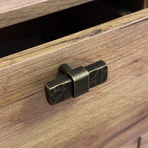 May include: Close-up of a bronze-toned drawer pull with a textured, rectangular design. The pull is mounted on a wooden drawer, with the drawer partially open, revealing a dark interior. The hardware has a rustic, industrial aesthetic.
