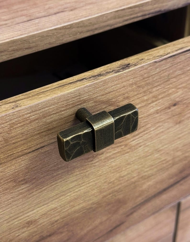 May include: Close-up of a bronze-toned drawer pull with a textured, rectangular design. The pull is mounted on a wooden drawer, with the drawer partially open, revealing a dark interior. The hardware has a rustic, industrial aesthetic.