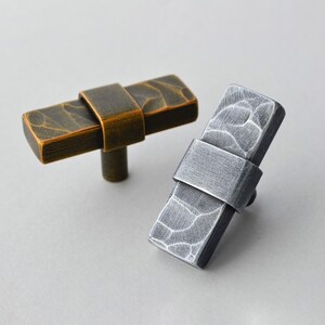 May include: Two rectangular cabinet pulls. One is bronze-colored with a textured surface and a curved band. The other is silver-colored with a similar texture and band. Both have a cylindrical stem.