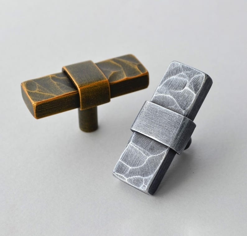 May include: Two rectangular cabinet pulls. One is bronze-colored with a textured surface and a curved band. The other is silver-colored with a similar texture and band. Both have a cylindrical stem.