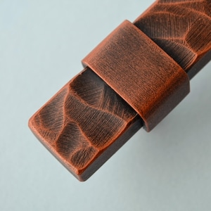 May include: Close-up of a copper-colored cabinet pull with a textured, carved design. The pull features a rectangular base with a strap-like handle. The finish has a rustic, aged appearance, suggesting a vintage or handcrafted aesthetic.