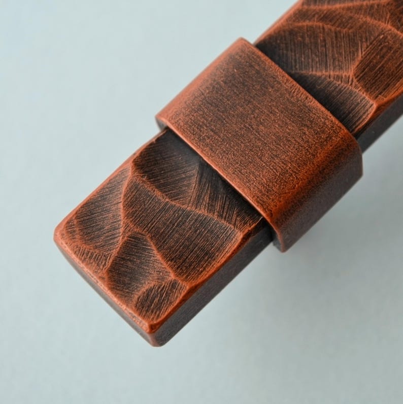 May include: Close-up of a copper-colored cabinet pull with a textured, carved design. The pull features a rectangular base with a strap-like handle. The finish has a rustic, aged appearance, suggesting a vintage or handcrafted aesthetic.