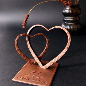 K&ouml;nnte beinhalten: Eine kupferfarbene Metallskulptur aus zwei ineinander verschlungenen Herzen auf einem quadratischen Sockel. Der Sockel ist mit dem Text "HI! II 05.06.2016 HENRY & ANNA" graviert.