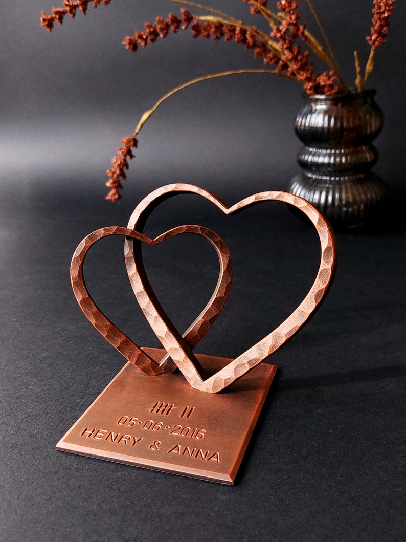 K&ouml;nnte beinhalten: Eine kupferfarbene Metallskulptur aus zwei ineinander verschlungenen Herzen auf einem quadratischen Sockel. Der Sockel ist mit dem Text "HI! II 05.06.2016 HENRY & ANNA" graviert.