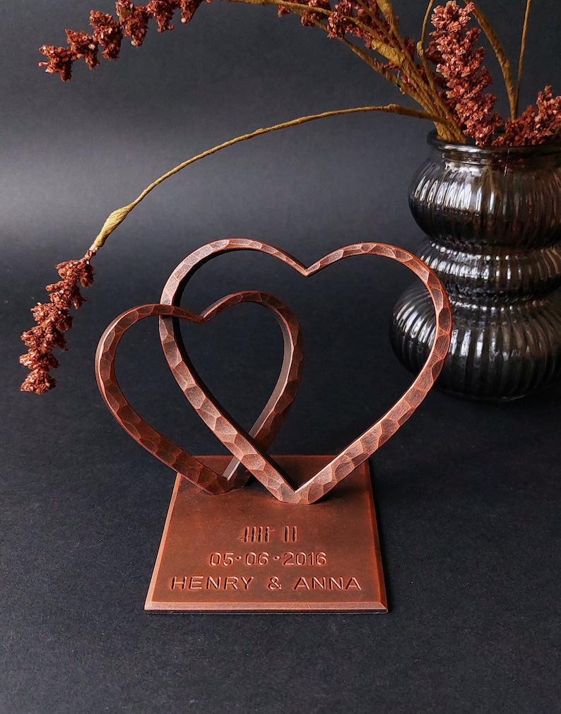 K&ouml;nnte beinhalten: Eine kupferfarbene Metallskulptur aus zwei ineinander verschlungenen Herzen auf einem quadratischen Sockel. Der Sockel ist mit dem Datum "05.06.2016" und den Namen "HENRY & ANNA" graviert.