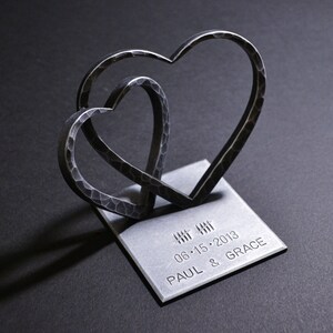 May include: Two interlocking metal hearts on a silver base with the inscription "06.15.2013 PAUL & GRACE".