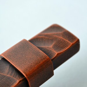 May include: Close-up of a rectangular, copper-colored wooden drawer pull with a textured surface. The pull has a carved design and a curved handle. The hardware is likely for a cabinet or drawer.