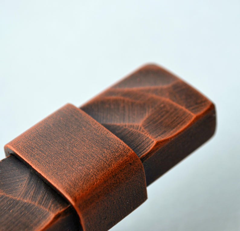 May include: Close-up of a rectangular, copper-colored wooden drawer pull with a textured surface. The pull has a carved design and a curved handle. The hardware is likely for a cabinet or drawer.