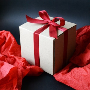 K&ouml;nnte beinhalten: Eine braune Geschenkbox aus Pappe mit einem roten Satinband, das zu einer Schleife gebunden ist. Die Box steht auf einem roten Seidenpapier-Hintergrund.
