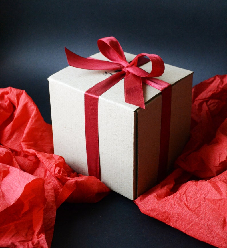 K&ouml;nnte beinhalten: Eine braune Geschenkbox aus Pappe mit einem roten Satinband, das zu einer Schleife gebunden ist. Die Box steht auf einem roten Seidenpapier-Hintergrund.