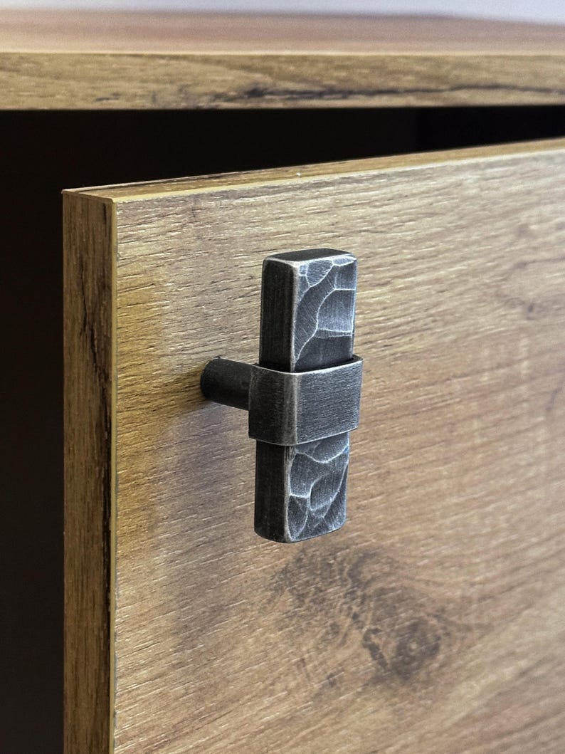 May include: A close-up of a cabinet door with a dark, textured metal handle. The handle is rectangular with a hammered appearance, mounted on a wooden cabinet with a warm, brown finish. The cabinet door is partially open.