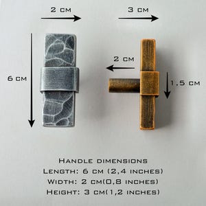 May include: Two cabinet handles are shown, one silver and one bronze. The silver handle is 6 cm (2.4 inches) long, 2 cm (0.8 inches) wide, and 3 cm (1.2 inches) high. The bronze handle is 6 cm (2.4 inches) long, 2 cm (0.8 inches) wide, and 1.5 cm (0.6 inches) high.