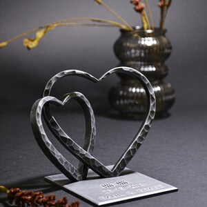May include: Two interlocking metal hearts on a square base with the inscription "08.08.2019 Paul & Grace".