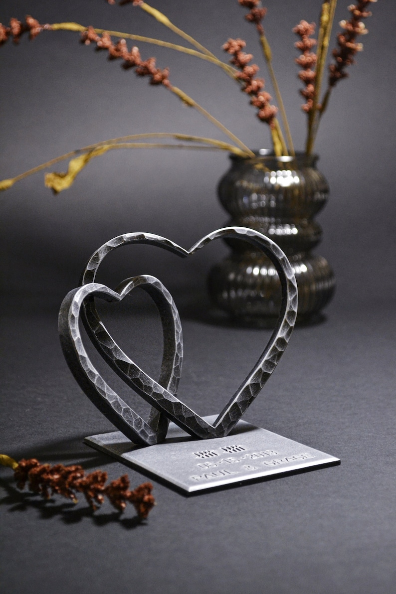 May include: Two interlocking metal hearts on a square base with the inscription "08.08.2019 Paul & Grace".