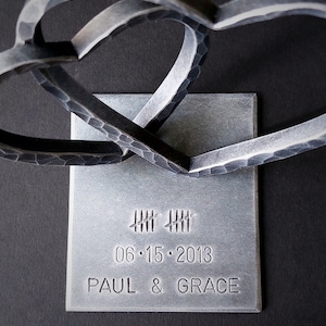 May include: Two intertwined metal hearts on a silver square base with the date 06.15.2013 and the names Paul & Grace engraved.