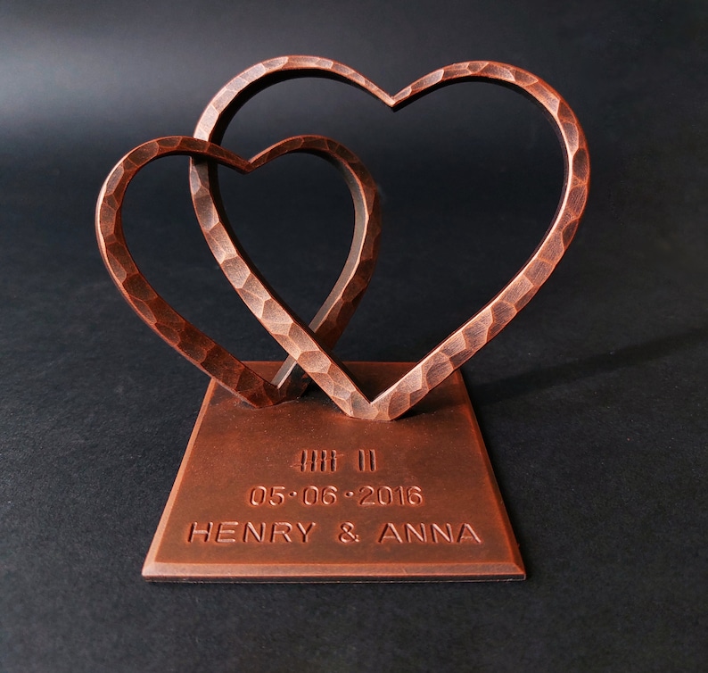 K&ouml;nnte beinhalten: Zwei ineinander verschlungene Kupferherzen auf einem Sockel mit dem Datum "05.06.2016" und den Namen "HENRY & ANNA" graviert.