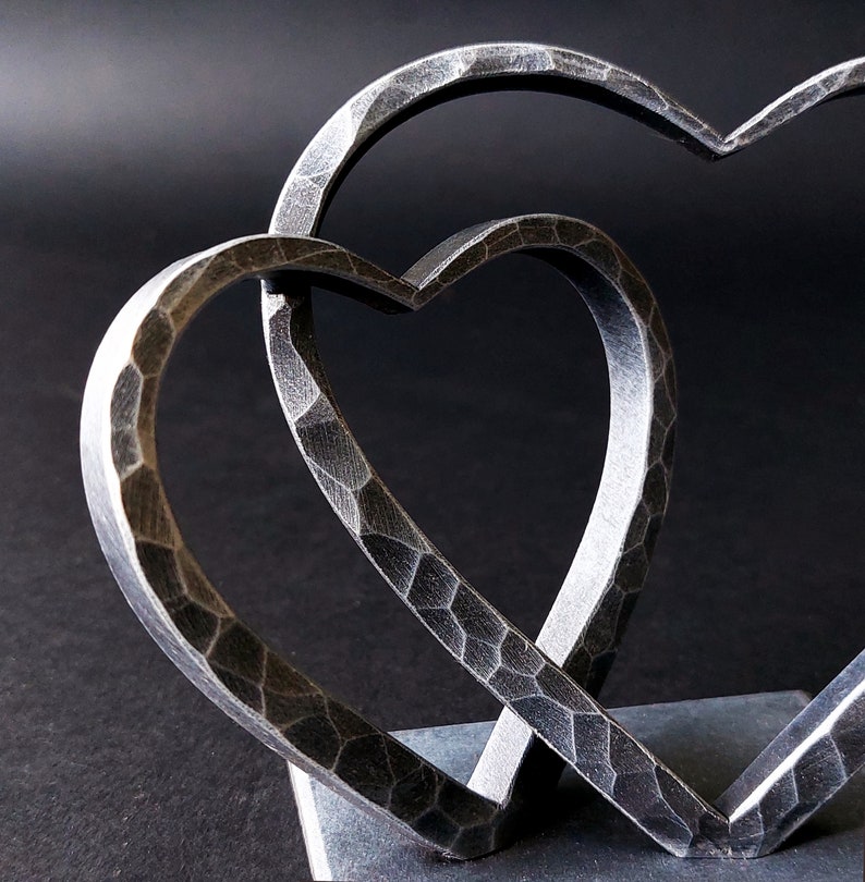 May include: Two interlocking metal hearts, each with a hammered texture, are joined at the base on a black slate surface.