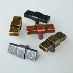 May include: A collection of five rectangular cabinet knobs in various metallic finishes. The knobs include bronze, black, copper, silver, and gold tones. Each knob features a textured surface and a central band detail, showcasing a modern design.