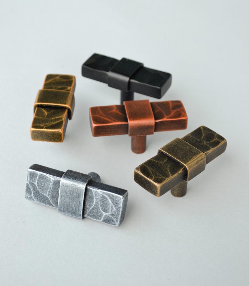 May include: A collection of five rectangular cabinet knobs in various metallic finishes. The knobs include bronze, black, copper, silver, and gold tones. Each knob features a textured surface and a central band detail, showcasing a modern design.