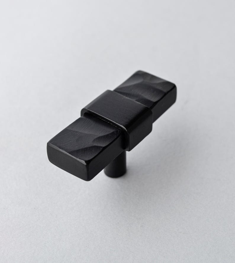 May include: A black cabinet knob with a rectangular shape and a textured surface. The knob has a central band and a stem for mounting. The hardware is made of metal and is suitable for furniture or cabinetry.