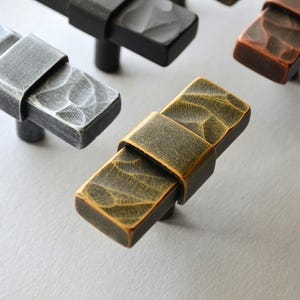 May include: Close-up of several rectangular cabinet pulls in various metallic finishes. The pulls have a textured surface and a band around the center. Finishes include silver, black, bronze, and a golden hue. These hardware pieces are designed for furniture.