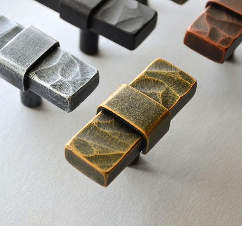 May include: Close-up of several rectangular cabinet pulls in various metallic finishes. The pulls have a textured surface and a band around the center. Finishes include silver, black, bronze, and a golden hue. These hardware pieces are designed for furniture.