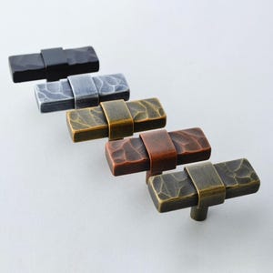 May include: A collection of rectangular cabinet knobs in various metallic finishes, including black, silver, bronze, copper, and antique brass. Each knob features a textured surface and a curved metal band. The knobs are arranged diagonally against a white background.