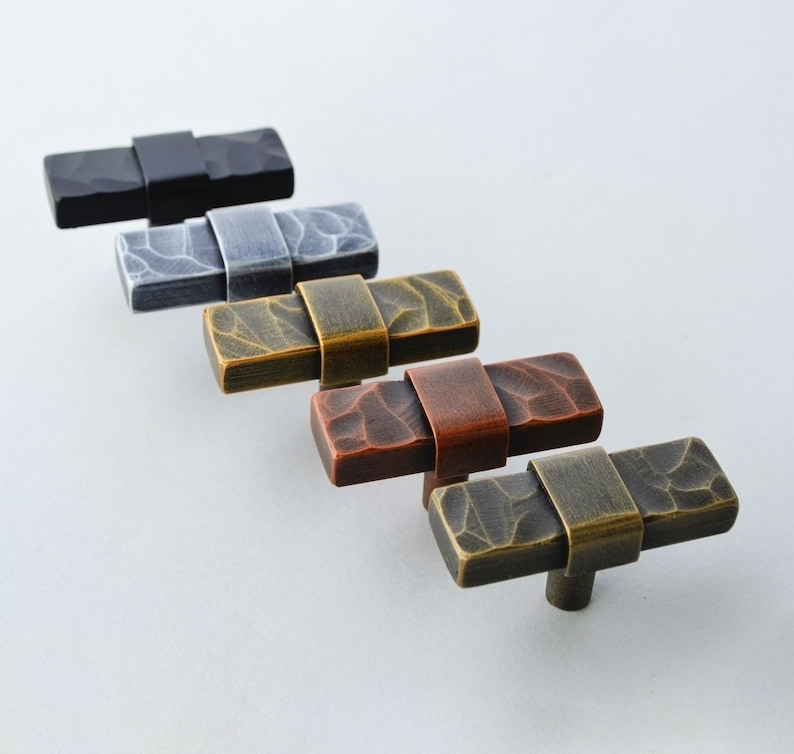 May include: A collection of rectangular cabinet knobs in various metallic finishes, including black, silver, bronze, copper, and antique brass. Each knob features a textured surface and a curved metal band. The knobs are arranged diagonally against a white background.