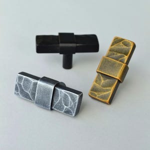 May include: Three decorative cabinet knobs in different finishes. One is black, one is silver, and one is gold. Each knob has a rectangular shape with a textured surface and a metal band around the center.