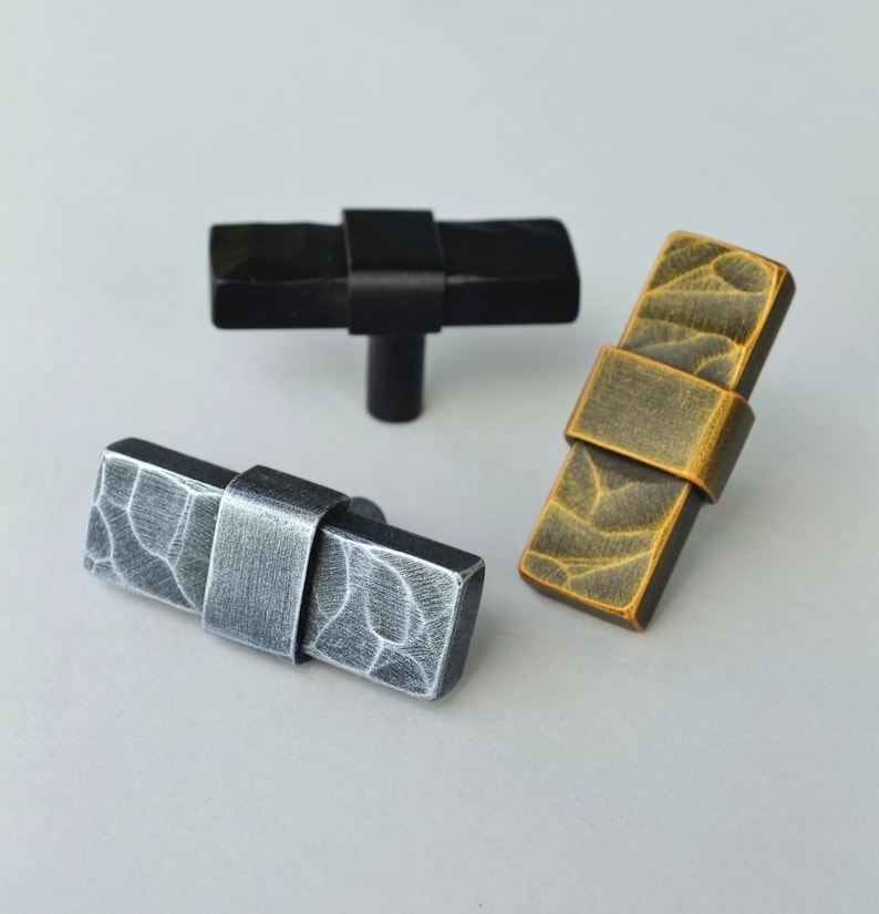 May include: Three decorative cabinet knobs in different finishes. One is black, one is silver, and one is gold. Each knob has a rectangular shape with a textured surface and a metal band around the center.