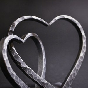 May include: Two interlocking metal hearts, each with a hammered texture, create a unique and modern design. The hearts are silver in color and are displayed against a dark background.