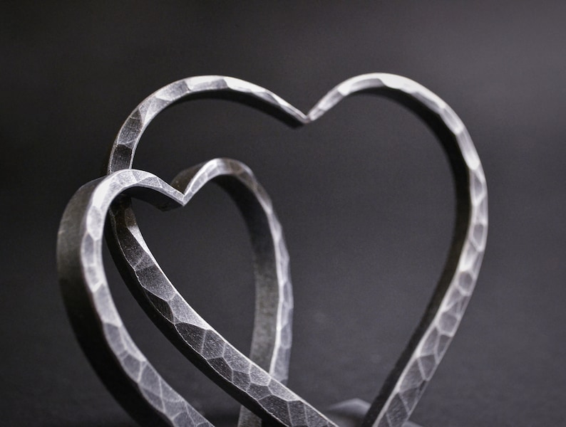 May include: Two interlocking metal hearts, each with a hammered texture, create a unique and modern design. The hearts are silver in color and are displayed against a dark background.