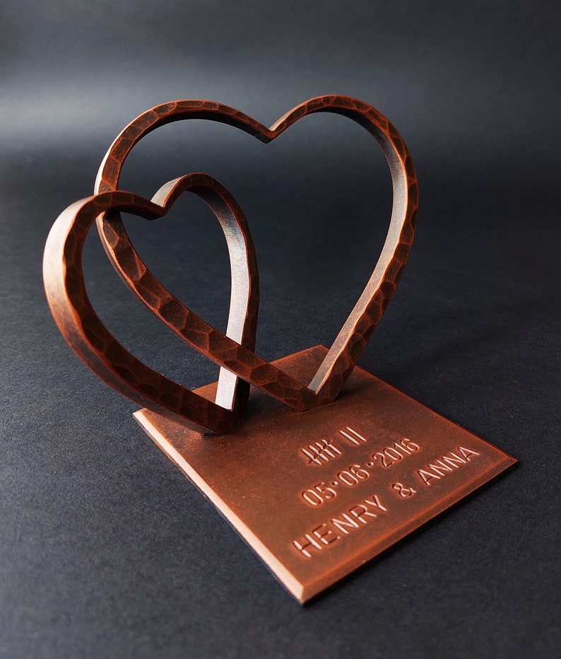 K&ouml;nnte beinhalten: Eine kupferfarbene Metallskulptur aus zwei ineinander verschlungenen Herzen auf einem quadratischen Sockel. Der Sockel ist mit dem Text "4.08.11 05.06.2016 HENRY & ANNA" graviert.