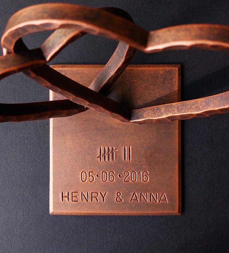 K&ouml;nnte beinhalten: Eine kupferfarbene Metallsculptur aus zwei ineinander verschlungenen Herzen mit einem rechteckigen Sockel. Der Sockel ist mit dem Datum "05.06.2016" und den Namen "HENRY & ANNA" graviert.
