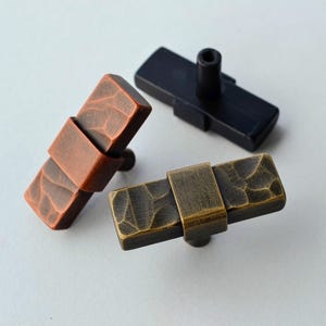May include: Three rectangular cabinet knobs in different finishes: copper, black, and antique brass. Each knob features a textured surface and a metal band detail. The knobs are designed for easy installation on furniture.