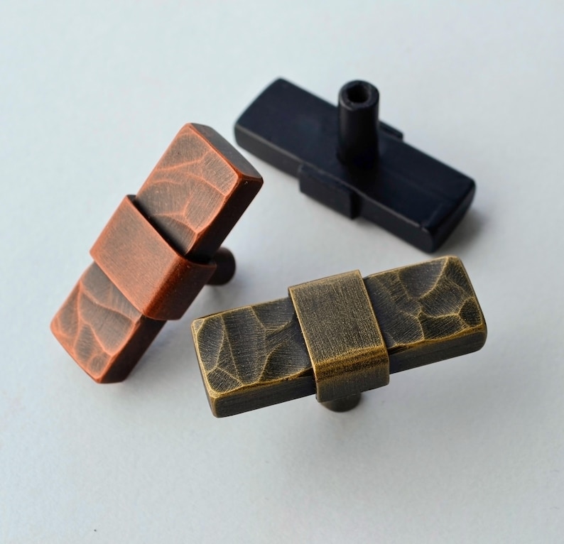 May include: Three rectangular cabinet knobs in different finishes: copper, black, and antique brass. Each knob features a textured surface and a metal band detail. The knobs are designed for easy installation on furniture.
