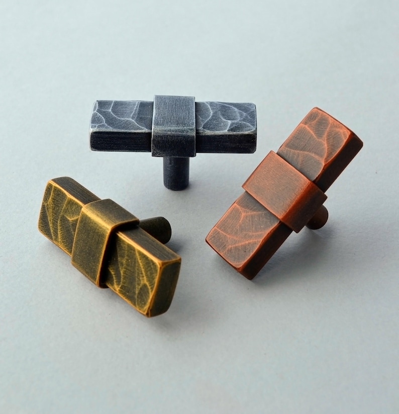 May include: Three rectangular cabinet knobs in bronze, black, and copper tones. Each knob features a textured surface and a central band detail. The knobs are designed for furniture and cabinet hardware.